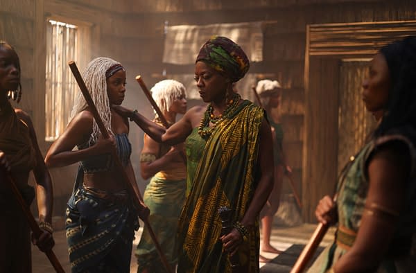 Thuso Mbedu as Zélie and Viola Davis as Mama Agba stand among a group of women in a scene from Children of Blood and Bone, holding staffs in a warm, rustic interior.
