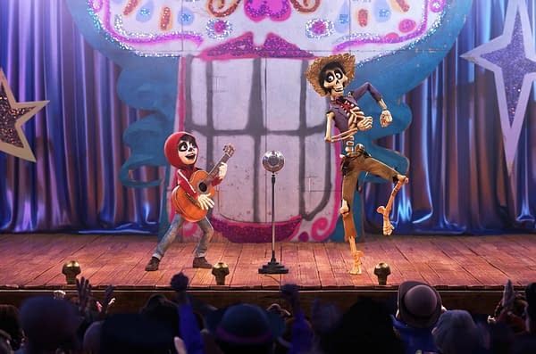 A scene from Coco featuring a young boy with a guitar and a skeleton in a hat performing on stage, with a colorful skull backdrop and an audience in front.