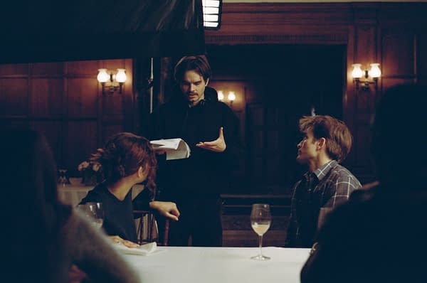 A behind-the-scenes moment during a film shoot, featuring a director in a black hoodie discussing a scene with two actors seated at a table, while a glass of wine sits in front of one actor amidst a dimly lit, elegant setting.