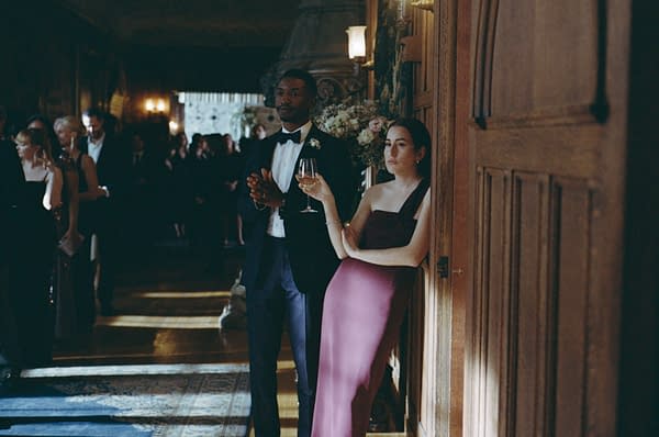 A man in a tuxedo and a woman in a burgundy gown stand together in a dimly lit room filled with party guests. The woman leans against a wall, holding a glass of drink, while the man stands beside her, lightly clapping.