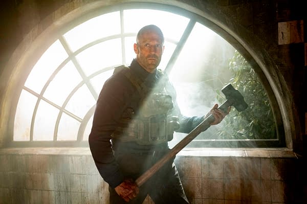 A muscular man stands in a smoky room, holding a heavy mallet and wearing tactical gear, with a large window behind him. The scene is dramatic, emphasizing the character Levon Cade from the film 'A WORKING MAN' directed by David Ayer.