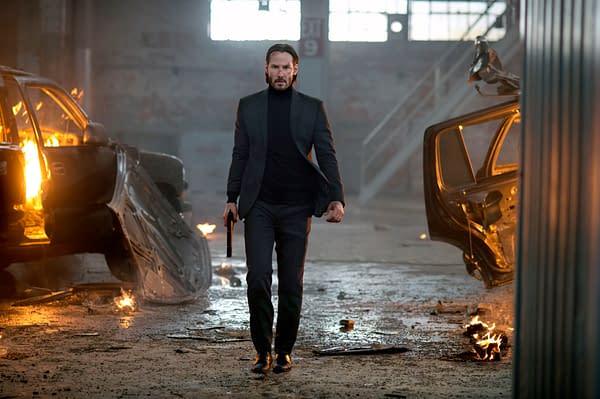 A man in a black suit and turtleneck walks through a gritty, dimly lit environment, holding a pistol, with flames erupting from overturned cars in the background.