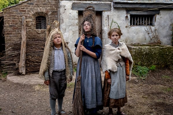 Three children dressed in historical costumes stand outside a rustic building. The girl in the center holds a broom, while the others wear unique head coverings and garments that reflect a bygone era, likely from the film 'Hamnet.'