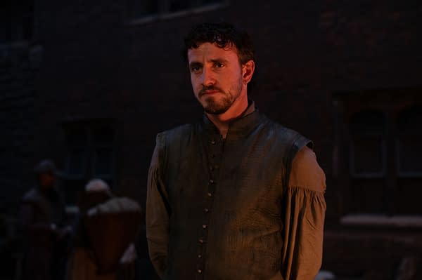 In the film 'Hamnet,' Paul Mescal portrays William Shakespeare, dressed in period attire with a somber expression, standing in a dimly lit setting that suggests a historical context.