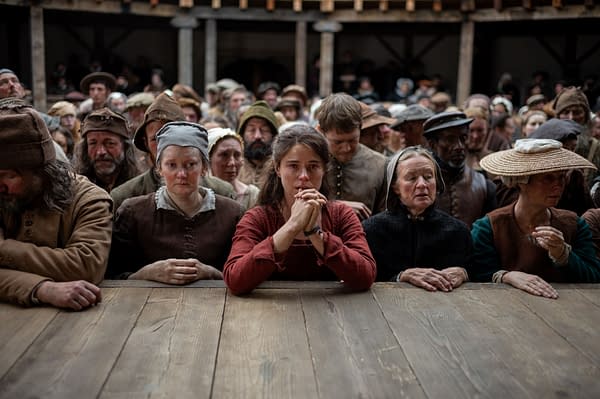 A still from the film 'Hamnet,' showcasing a group of somber characters in period attire, gathered in a crowded setting. Central to the image is a young woman with long dark hair, looking anxious as she grips her hands together at a wooden table, surrounded by others displaying a range of emotions.