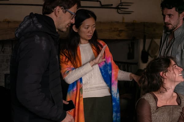 On the set of the film 'Hamnet', director Chloé Zhao, wearing a colorful tie-dye shawl, engages with actors Jessie Buckley and Paul Mescal, while a male crew member observes. The setting features a rustic backdrop with traditional tools visible.