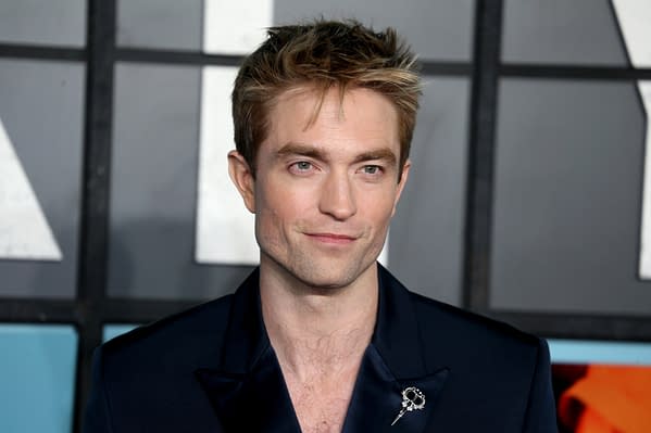 A close-up portrait of a man with light brown hair styled upward, wearing a dark suit with a decorative pin on his lapel. He has a subtle smile and is positioned against a blurred background of geometric shapes.