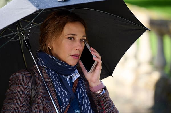 Rashida Jones holds a smartphone to her ear while standing under a black umbrella, dressed in a stylish, patterned jacket and scarf. She appears to be engaged in an important conversation.