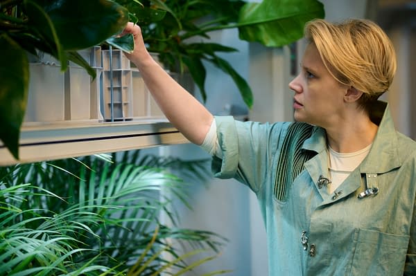 A scene from 'In The Blink Of An Eye' featuring a woman with short blonde hair, dressed in a green outfit, gently adjusting a plant while observing it closely. Lush foliage surrounds her, creating a vibrant indoor garden atmosphere.