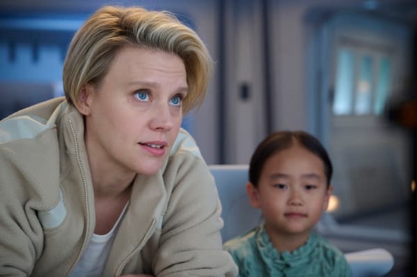 A still from the movie 'In The Blink Of An Eye' featuring two characters: an adult woman with short blonde hair and blue eyes, wearing a beige jacket, and a young girl with dark hair styled in two braids, wearing a green top. They are both gazing intently, suggesting a moment of connection or discovery.