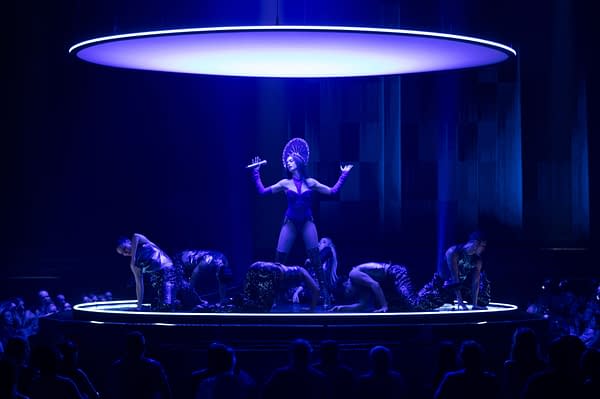 A theatrical performance featuring a singer in a sparkling purple outfit with dramatic gloves, surrounded by dancers on a circular stage illuminated by blue lighting. The audience can be seen watching intently.