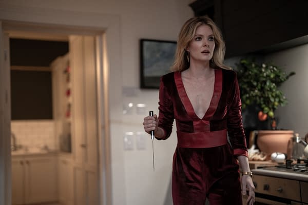 In the film 'Drop,' actress Meghann Fahy portrays Violet, holding a kitchen knife with a tense expression while wearing a deep red velvet jumpsuit in a domestic setting.