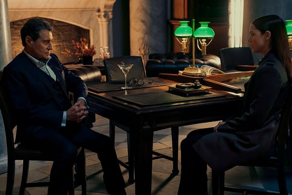 In a dimly lit room, Gabriel Byrne as The Chancellor sits across the table from Ana de Armas as Eve, both looking serious. A green lamp and a glass of martini add to the dramatic atmosphere.