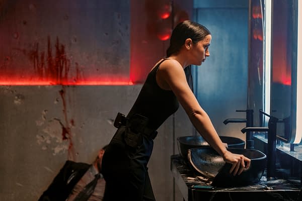 A woman in a black outfit leans over a black sink, her expression serious, in a dimly lit location with red accents and visible signs of distress. The scene hints at a tense moment in the film 'Ballerina,' featuring Ana de Armas as the character Eve.