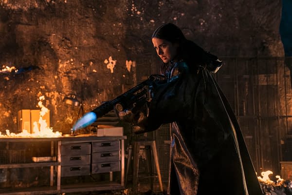A scene from 'Ballerina' featuring a woman in a dark, shiny raincoat aiming a weapon with flame at the end, surrounded by a dimly lit room with flames in the background.