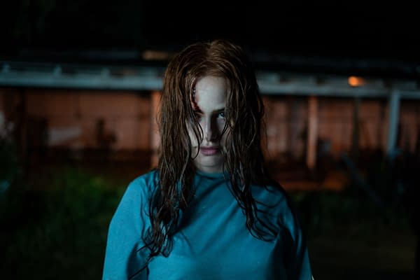 A woman with disheveled hair and an intense expression stands in a dimly lit outdoor setting, wearing a blue shirt. A small wound is visible on her forehead, adding to the eerie atmosphere.