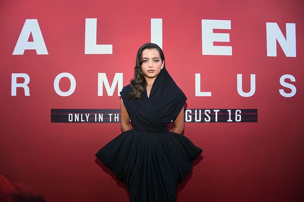 A woman in a dramatic black gown poses in front of a promotional backdrop featuring the title 'ALIEN: ROMULUS'. The backdrop is bright red, emphasizing the film release date.