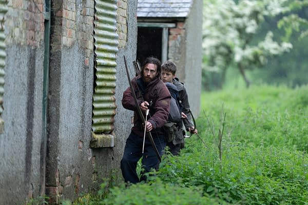 A tense scene showing two survivors, one with a bow and arrows and the other carrying a backpack, stealthily moving past a dilapidated building in a lush green environment, conveying a sense of urgency and survival.