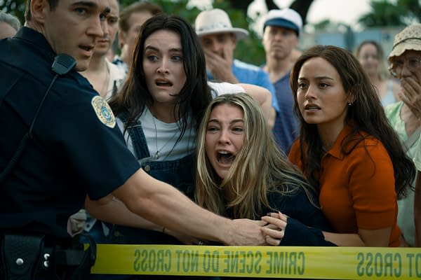 A tense scene featuring a police officer holding back a crowd, while three women display fear and distress, particularly one who is crying out. Yellow crime scene tape is visible in the foreground, contributing to the dramatic atmosphere.