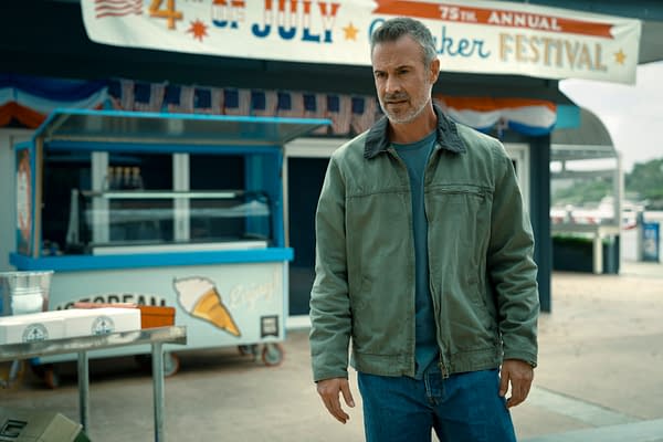 A still from the trailer of 'I Know What You Did Last Summer' featuring a man in a green jacket with gray hair, standing in front of an ice cream stand at a festive outdoor setting. The background displays a banner for a summer festival.