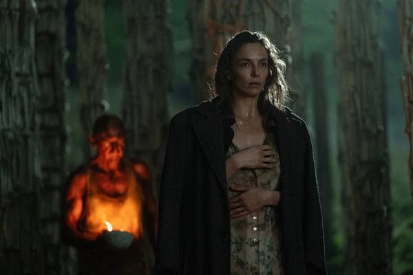 A dramatic scene featuring a woman with tousled hair wearing a floral dress and a long coat, holding her hand over her chest in a forest setting. In the background, a shadowy figure holds a lit candle, adding an eerie atmosphere.