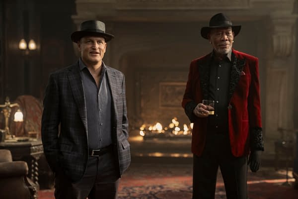 Woody Harrelson, dressed in a plaid suit and a black fedora, stands beside Morgan Freeman, who wears a red velvet jacket. They are in a dimly lit room with a cozy fireplace in the background, suggesting an atmosphere of intrigue.