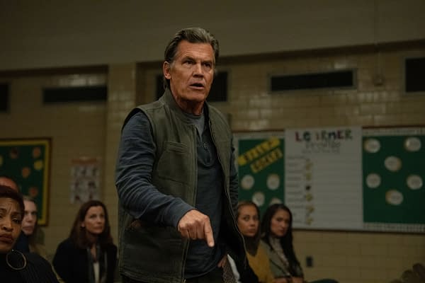 A man with gray hair and a serious expression stands before an audience, gesturing with his hand, in a brightly lit indoor setting. The background features a colorful bulletin board with visible educational materials.