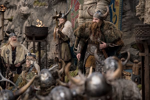 A scene from the live-action film 'How to Train Your Dragon,' featuring Stoick, portrayed by Gerard Butler, alongside other Viking characters in elaborate costumes with horns and fur, set against a rocky backdrop with torches and banners.