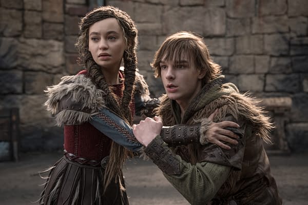 A scene from 'How To Train Your Dragon' featuring two characters, a young woman with braided hair in a red and grey costume, and a young man with tousled hair in a green hoodie and fur-trimmed clothing, looking distressed while holding onto each other in a medieval setting.