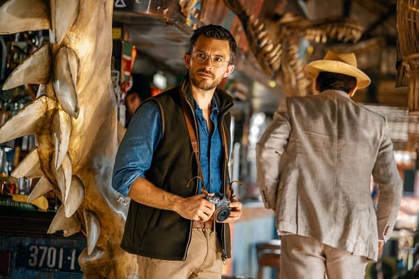 Jonathan Bailey portrays paleontologist Dr. Henry Loomis in a scene from 'Jurassic World Rebirth,' featuring him holding a camera and standing in front of a dinosaur skull in a vibrant setting.