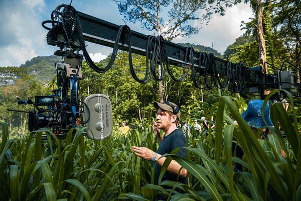 On the set of 'Jurassic World Rebirth,' director Gareth Edwards appears engaged while surrounded by tall green grass and a crew. A camera on a crane is positioned above, capturing the action in a lush outdoor environment.