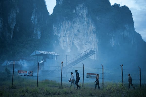 A scene from 'Jurassic World Rebirth' shows four characters walking along a foggy, overgrown area near a chain-link fence. In the background, a dilapidated building and rocky cliffs are visible, creating an eerie atmosphere.