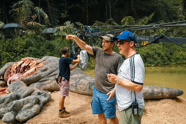 On the set of 'Jurassic World Rebirth,' director Gareth Edwards points while discussing a scene, flanked by crew members and a large, dinosaur-like prop in a lush, green environment.