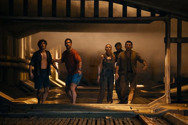 Group of five characters from 'Jurassic World Rebirth' standing in a dimly lit, industrial setting looking tense and alert, with a mix of expressions suggesting danger. The characters include Xavier Dobbs, Reuben Delgado, Zora Bennett, Duncan Kincaid, and Dr. Henry Loomis.