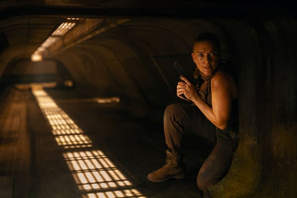 A woman crouches in a dimly lit environment, holding a handgun. She appears alert and focused, suggesting a scene of suspense from 'JURASSIC WORLD REBIRTH.'