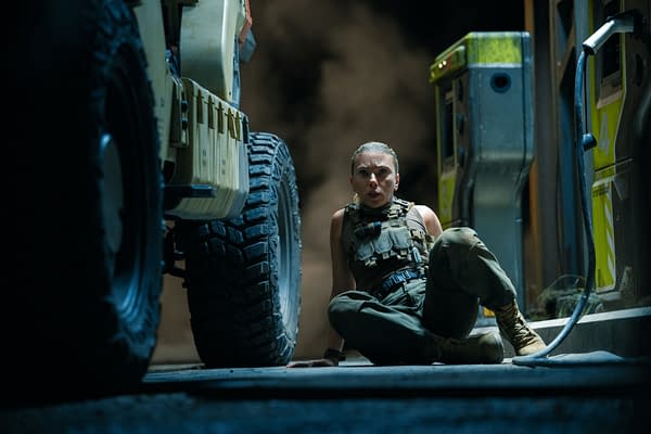 In a dark setting, a female character, Zora Bennett, portrayed by Scarlett Johansson, is crouched on the ground near a vehicle, exuding a sense of urgency and focus amid a tense moment from 'Jurassic World Rebirth.'