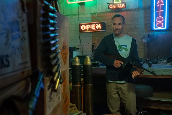 Bob Odenkirk portrays Hutch Mansell holding a gun in a dimly lit room featuring neon signs, surrounded by tools and military artifacts, in a scene from the film Nobody 2.