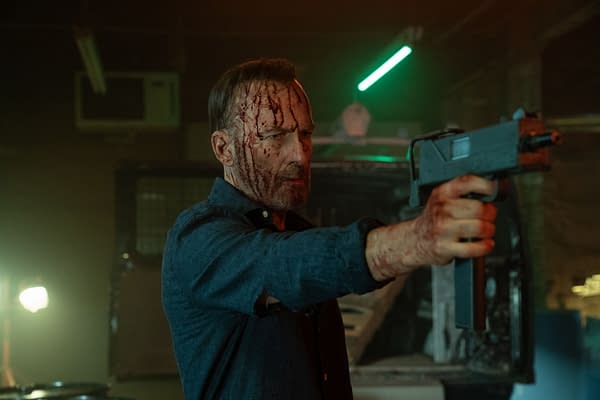 A man with a bloodied face and hands aims a gun in a dimly lit, industrial setting. The atmosphere is tense, highlighted by a green neon light.