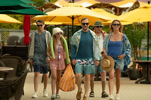 A scene from Nobody 2 featuring a group of five people walking together at a leisure location, each dressed in colorful summer attire and sunglasses, carrying various items like a bag and a hat, with yellow umbrellas visible in the background.