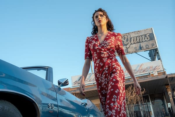 Margaret Qualley walks confidently past a vintage blue car, wearing a red floral dress, with a dilapidated shop sign in the background that reads "Yums Bakery." This scene is from Ethan Coen's film "HONEY DON'T!".