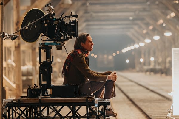 Wes Anderson sits on set with a film camera on a tripod in the foreground, surrounded by a dimly lit railway line that features overhead lights, during the production of 'The Phoenician Scheme.'