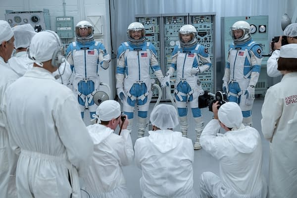 Four astronauts in blue and white space suits stand in a futuristic control room, surrounded by technicians in white protective clothing. The scene captures a moment of preparation and excitement for space travel.