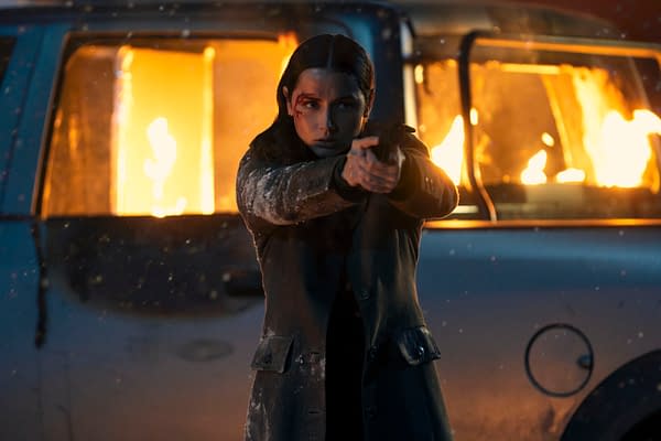 A scene from the movie 'Ballerina' featuring a woman, portrayed by Ana de Armas, pointing a gun while wearing a dark coat. Behind her, a vehicle is engulfed in flames.