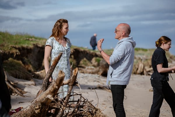 A behind-the-scenes moment on a seaside set, featuring a woman in a floral dress engaging in conversation with a man wearing a gray hoodie. A crew member is seen in the background, alongside driftwood scattered on the beach.