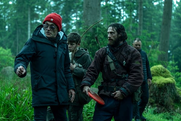 A group of four people in a forest, with one wearing a red beanie and gesturing, while another holds an arrow and bow. The scene conveys a sense of urgency and focus, suitable for a narrative about survival or adventure.