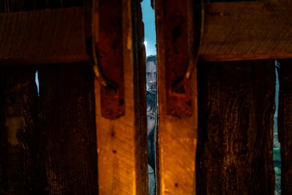 A cinematic scene viewed through a crack in wooden planks, revealing two faces with a tense expression. The atmosphere is moody and suspenseful, suggesting a moment of anticipation.
