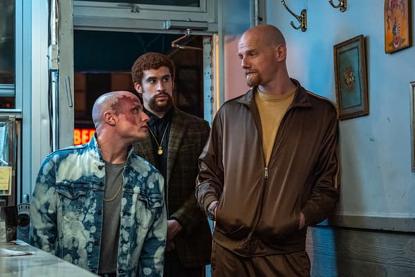Three men are in a dimly lit setting, with one man wearing a light blue denim jacket showing a facial injury, while another wears a dark plaid coat and the third is dressed in a brown tracksuit. The mood suggests tension or confrontation.