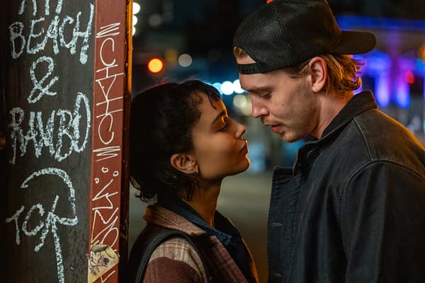 A romantic moment between two young adults, standing close and gazing into each other's eyes, with a graffiti-covered pole in the background. The scene is softly lit, suggesting a nighttime urban setting.