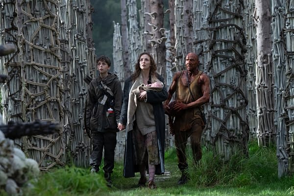 A scene from the post '28 Years Later:' featuring a boy holding hands with a woman who carries a baby, accompanied by a muscular man, all walking through a dense forest of tall, unique trees with bark resembling bones.