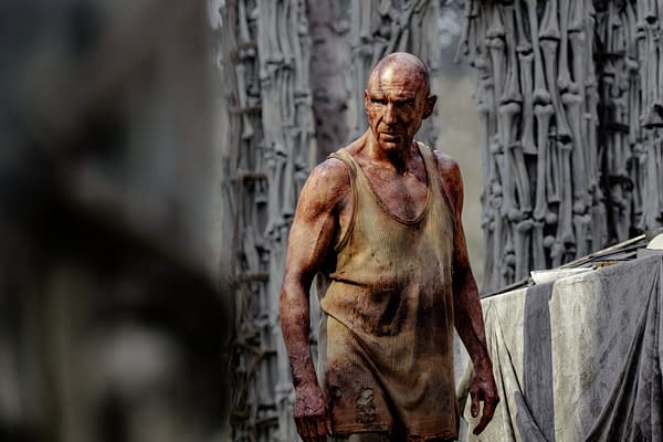 A rugged, muscular man in a torn tan tank top is standing amidst a desolate, eerie environment, with muddy skin and a serious expression. The background features skeletal structures and a stark, dark atmosphere.